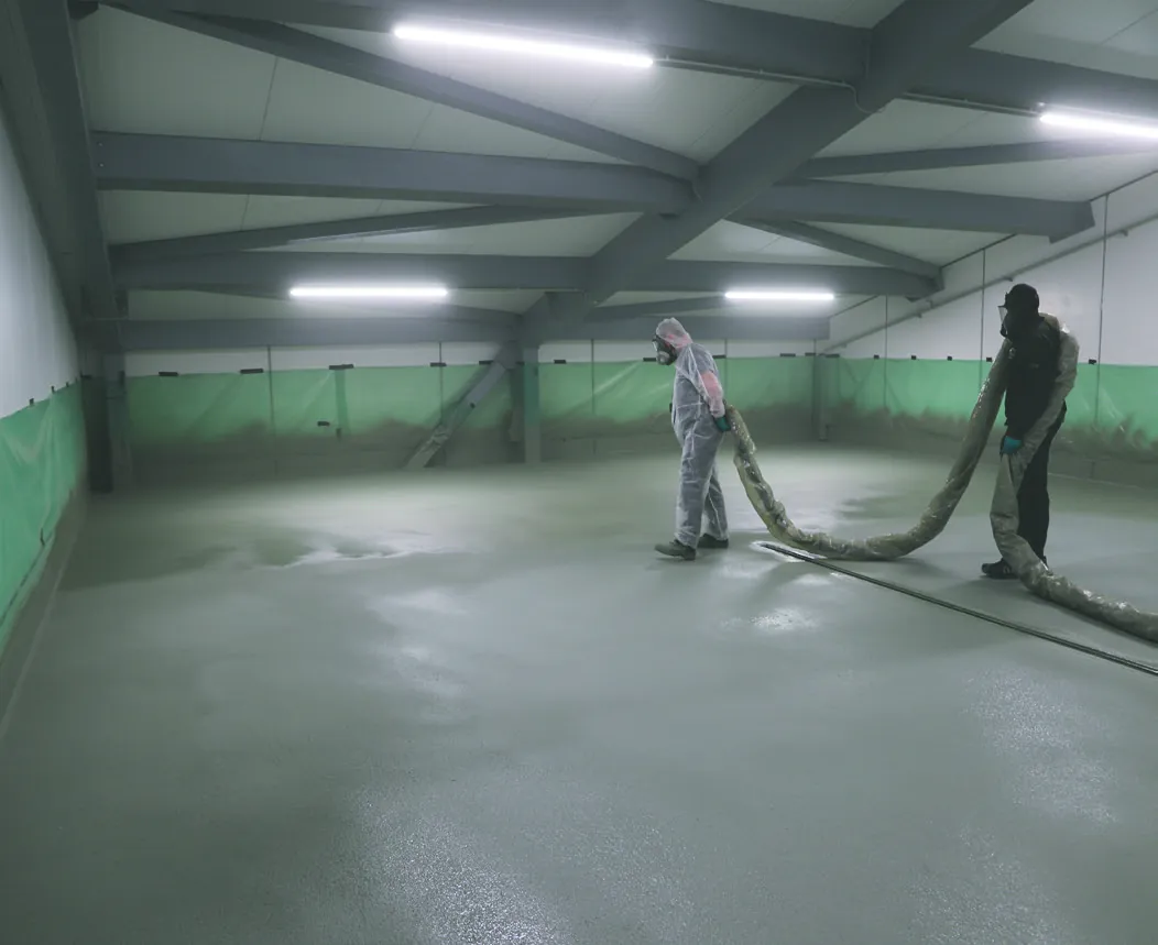 Two applicators walking the spray hose across a wide hall floor at MOWI, the fresh BAS Flexcoat membrane wet behind them.