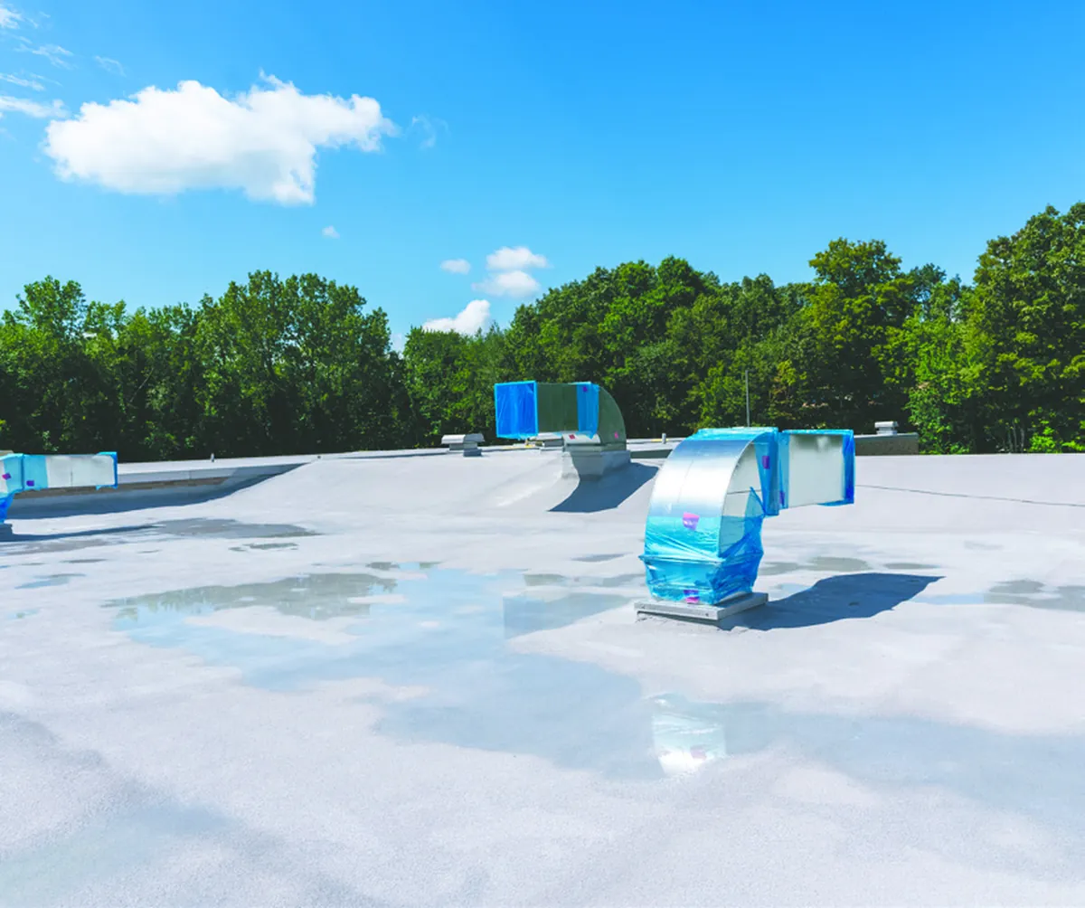 A flat industrial roof coated in pale BAS Flexcoat, with rooftop units and a line of trees beyond.